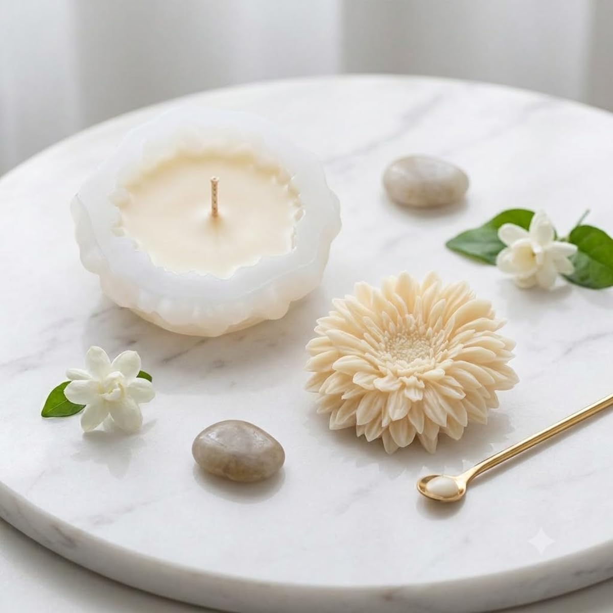 A styled product shot featuring a white wax candle in a textured, flower-like holder alongside decorative elements including a cream chrysanthemum flower, white jasmine blossoms, smooth stones, and a gold spoon on a white marble tray.