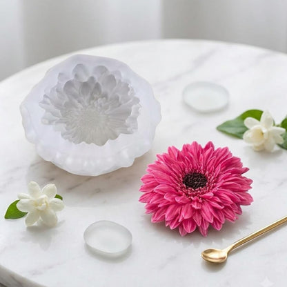 A white silicone mold with a detailed flower design, shown alongside a matching pink gerbera daisy flower and small white flowers on a white marble surface.