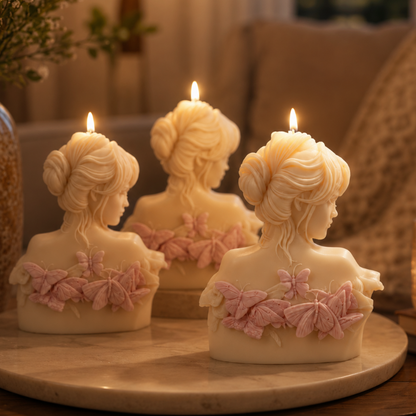 Three lit cream-colored pillar candles shaped like female busts with pink butterfly and floral accents, displayed on a round marble tray in a cozy indoor setting.
