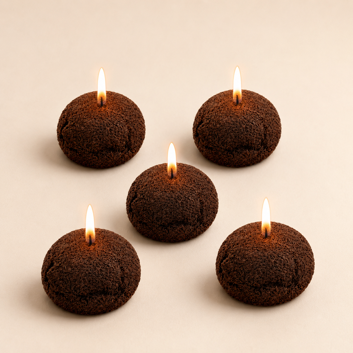 Five small dark brown textured candles with lit flames, arranged on a plain warm beige background.