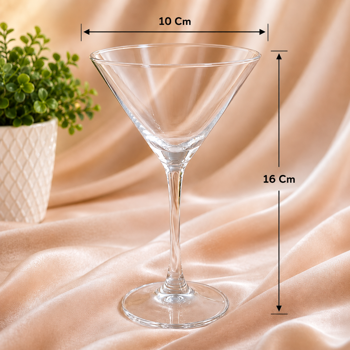 A clear martini-style glass on draped beige fabric with overlaid dimension arrows and labels showing a width of 10 cm and a height of 16 cm. A small potted green plant appears at the left edge.