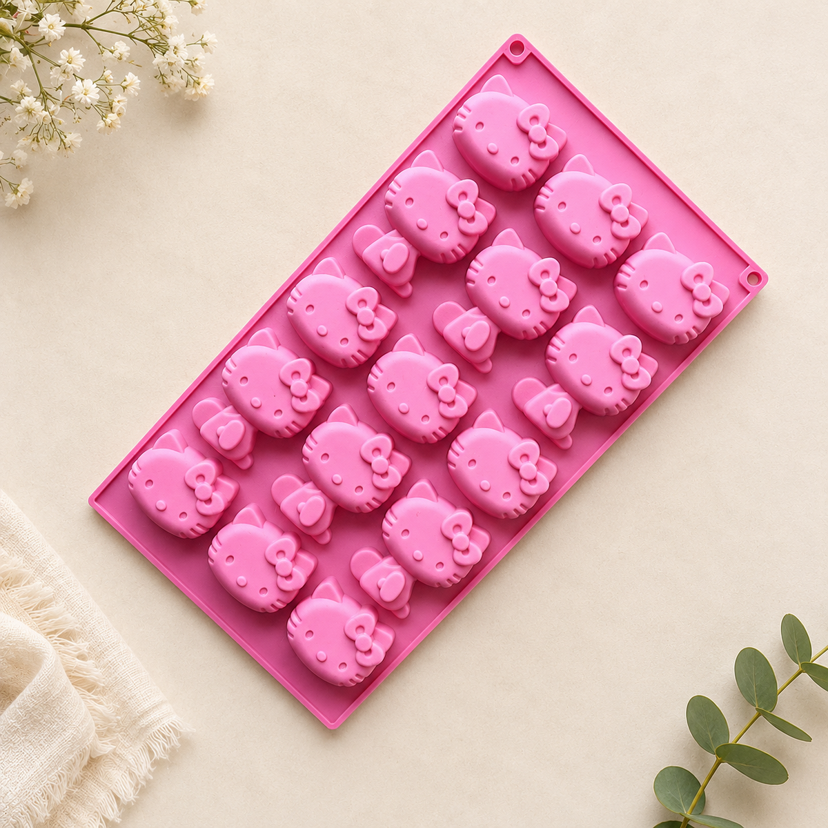 A pink rectangular silicone mold with multiple Hello Kitty face cavities, photographed on a light beige surface with small white flowers and green leaves as styling props.