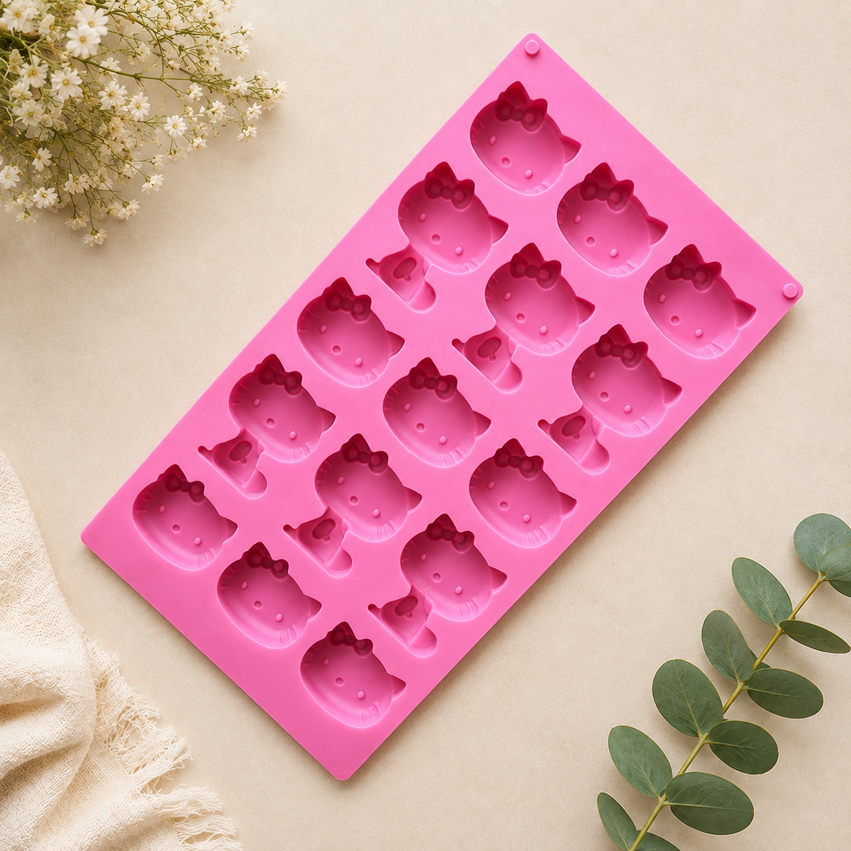 A pink silicone mold tray with multiple Hello Kitty face cavities, photographed on a beige surface with small white flowers and green leaves as styled props.
