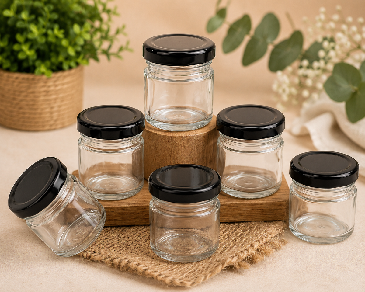 Five clear glass jars with black screw-on lids arranged on a wooden block and burlap fabric, with decorative plants in the background.