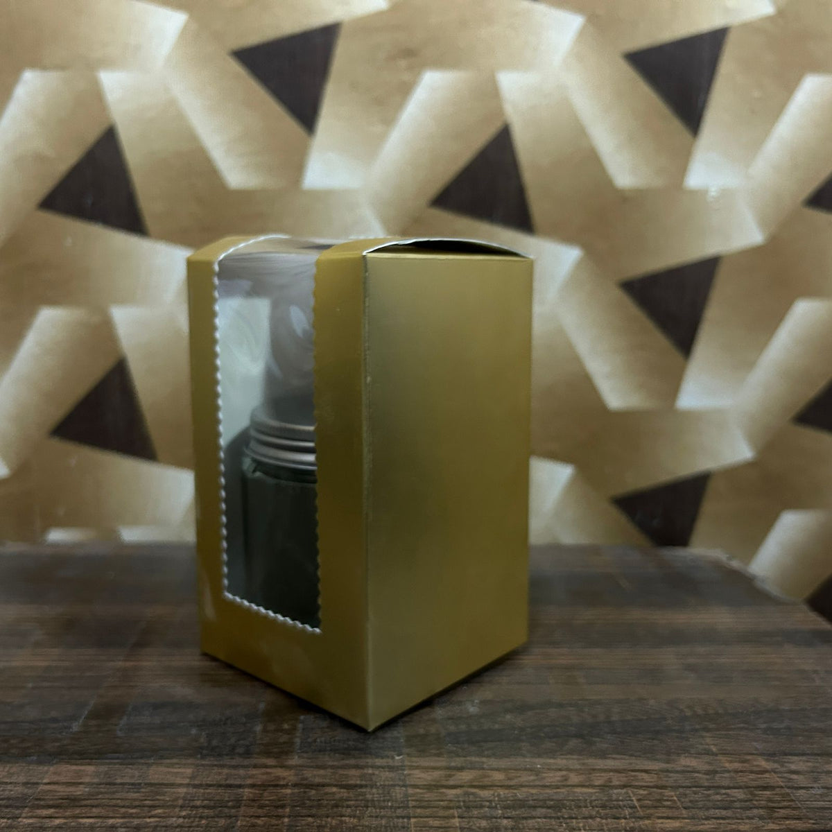 Yellow box with a clear window containing a black cylindrical object on a textured surface with geometric patterned background.