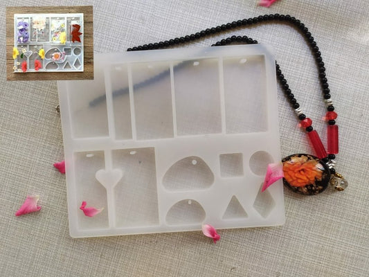 A silicone resin mold in the shape of a pendant with various cut-out designs, accompanied by a red beaded necklace and a few scattered flower petals on a beige fabric surface.