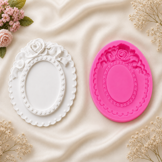 A white decorative oval frame mold and a matching bright pink oval silicone mold, displayed on a cream satin fabric with small pink flowers and baby's breath props.
