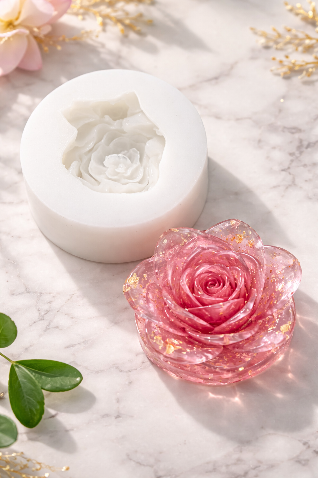 Pink rose-shaped soap next to a white rose mold on a marble surface with flowers and leaves.