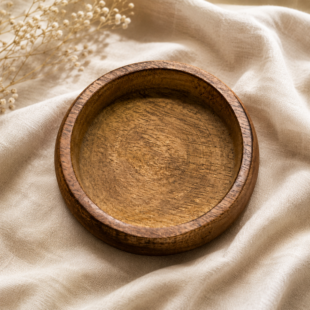 A round wooden tray with a smooth, polished finish and visible wood grain, resting on a draped beige linen fabric with dried baby's breath flowers in the corner.